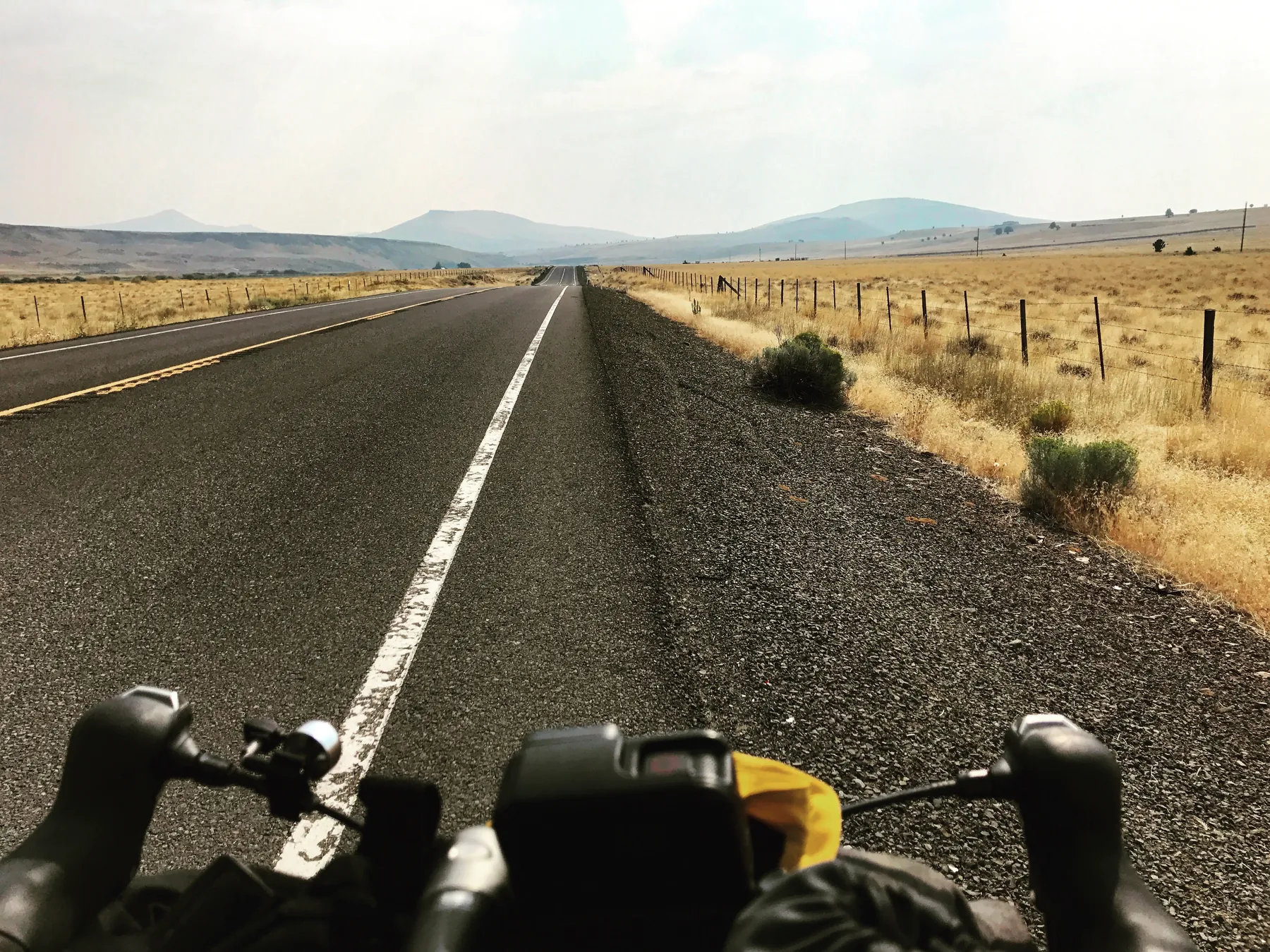 A long empty road stretching through dry open countryside, seen from bicycle handlebars.